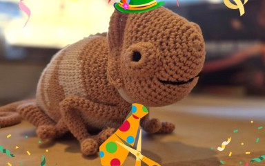 Gehäkelter Dinosaurier mit Partyhut und bunter Krawatte, umgeben von Konfetti.