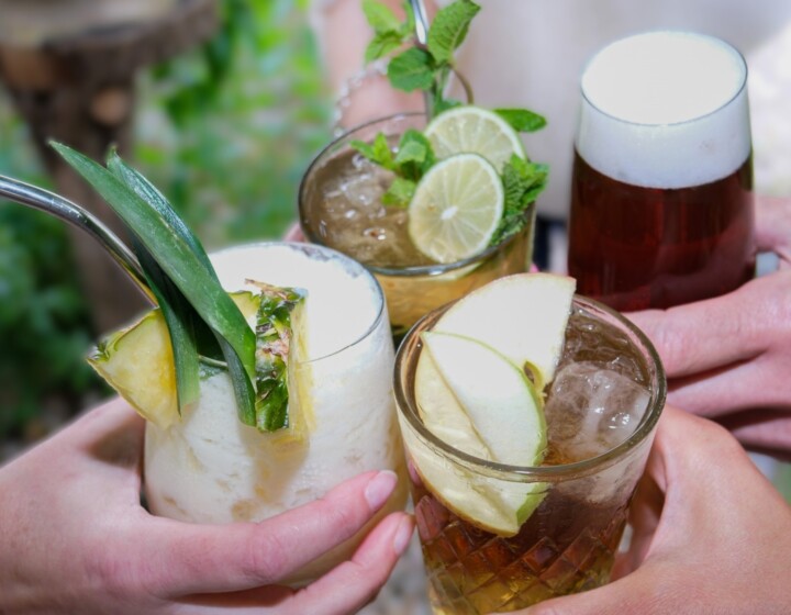 Vier Hände stoßen draußen mit Cocktails und Bier an, dekoriert mit Obst und Minze.