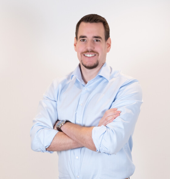 Lächelnder Mann mit Bart im hellblauen Hemd, Arme verschränkt, vor weißem Hintergrund.