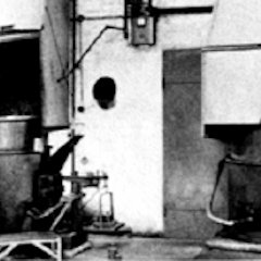 Schwarzweißfoto einer Werkhalle mit großen Maschinen, Rohren, Kabeln und industriellen Geräten.