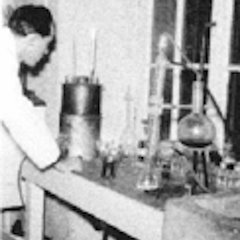 Wissenschaftler im weißen Kittel arbeitet an Labortisch mit Glasapparaturen und Messgeräten.