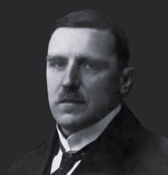 Schwarzweißporträt eines Mannes mit Schnurrbart im Anzug vor dunklem Hintergrund.