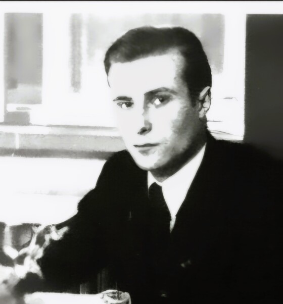 Schwarzweißporträt eines jungen Mannes im Anzug, ernst blickend, in Innenraum sitzend.