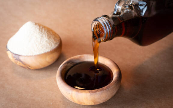 Dunkler Sirup wird aus Glasflasche in Holzschale gegossen, daneben Schale Zucker.
