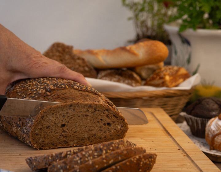 Hand schneidet ein Körnerbrot auf Holzbrett, daneben Korb mit Brötchen und Gebäck.