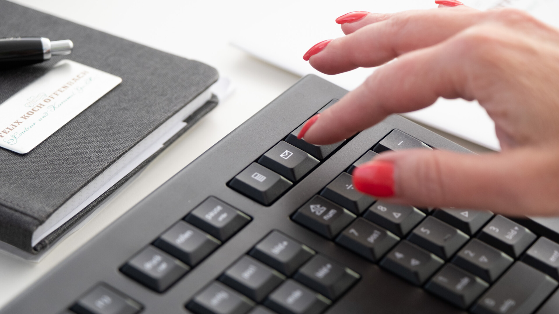 Hand mit roten Nägeln drückt Taste auf schwarzer Tastatur neben Notizbuch und Stift.