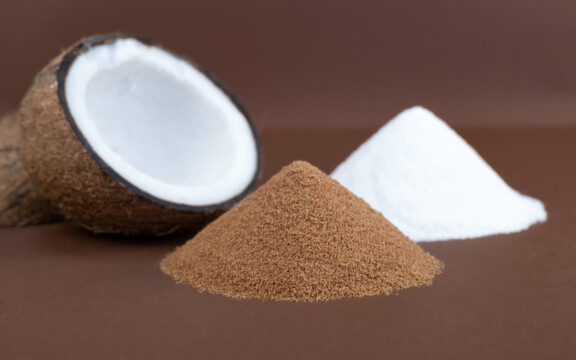 Halbe Kokosnuss neben braunem und weißem Pulverhaufen auf braunem Hintergrund arrangiert.