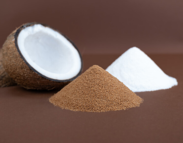 Halbe Kokosnuss neben braunem und weißem Pulverhaufen auf braunem Hintergrund arrangiert.