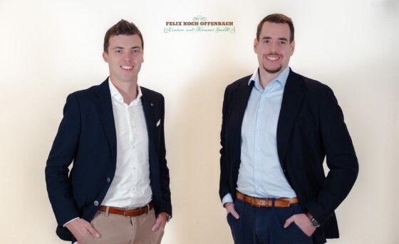 Zwei lächelnde Männer in Anzügen posieren vor hellem Hintergrund mit Firmenlogo oben.