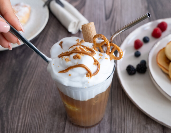 Hand rührt Sahnekaffee mit Metallstrohhalmen, daneben Pancakes und Beeren auf Tellern.