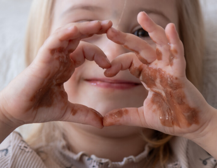 Lächelndes Kind formt Herz mit schokoladenverschmierten Händen und blickt in Kamera.