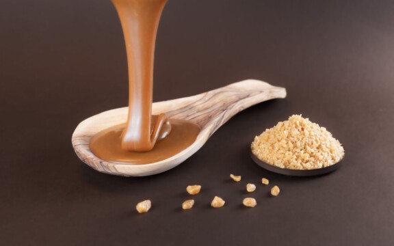 Flüssiges Karamell fließt in Holzlöffel neben Schale mit gehackten Nüssen, dunkler Hintergrund.