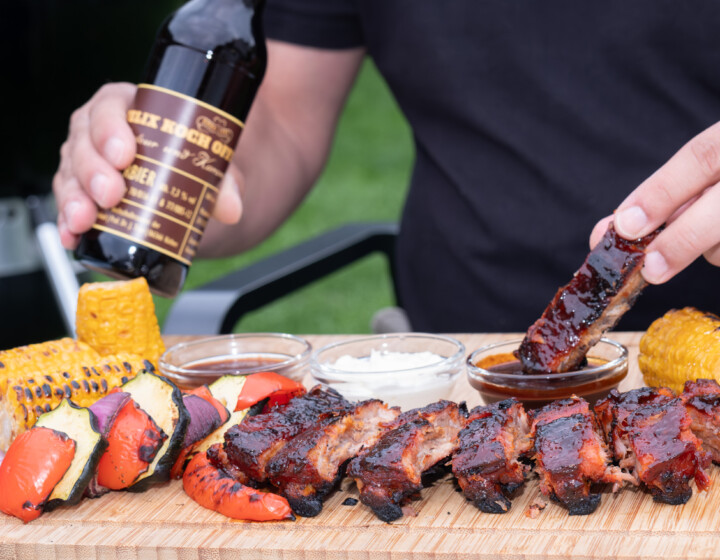 Person mariniert gegrillte Rippchen mit Bier, Grillgemüse, Maiskolben und Dips auf Holzbrett.