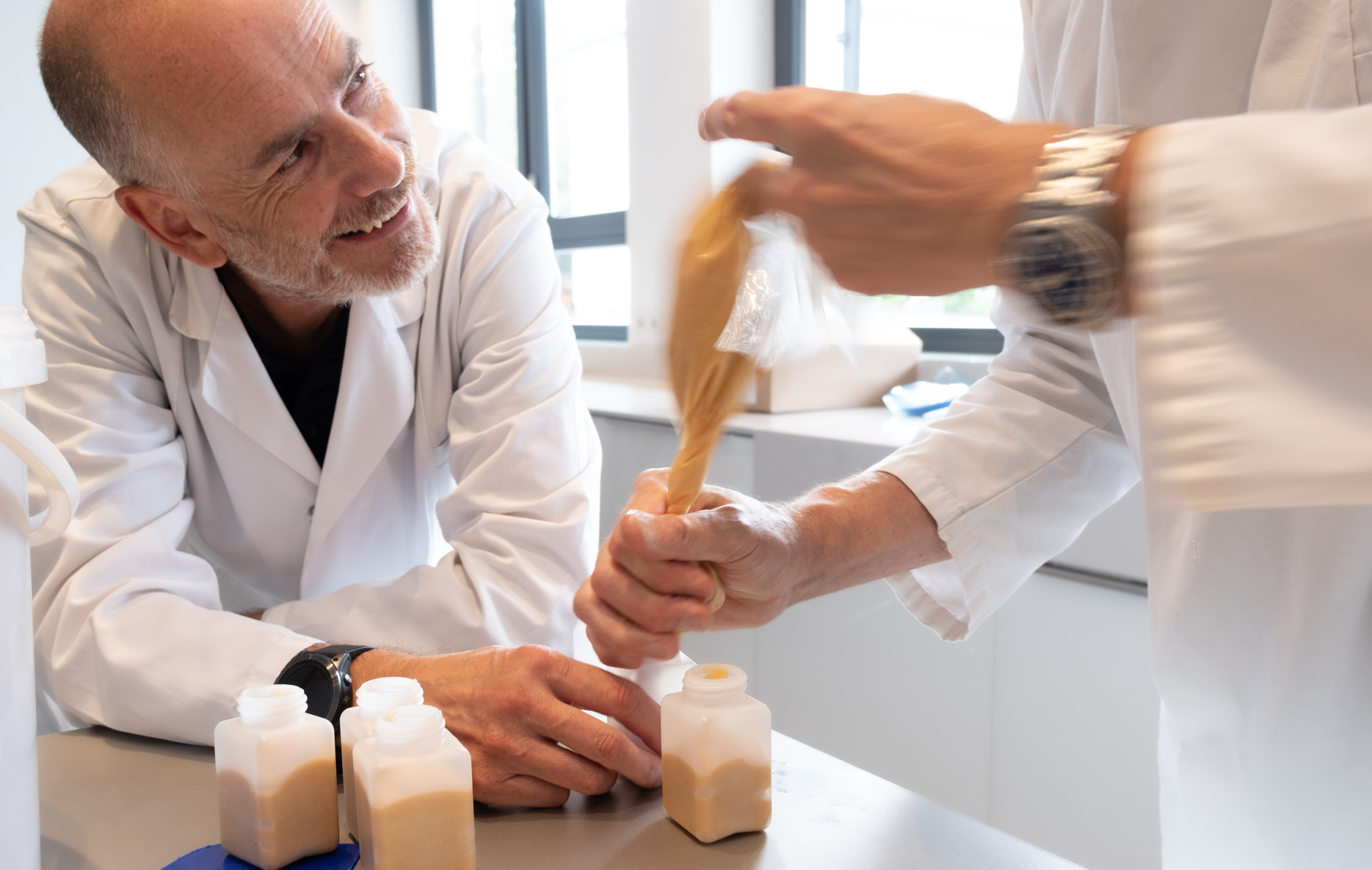 Zwei Laborpersonen füllen eine beige Flüssigkeit lachend in kleine Kunststofffläschchen ab.